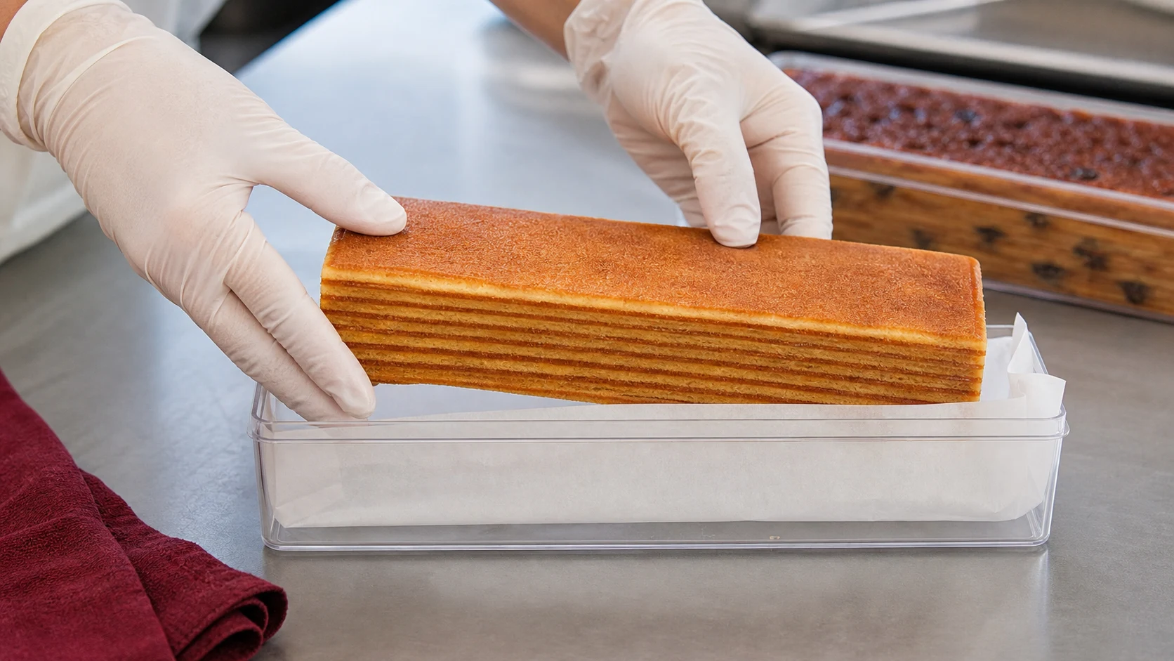 Gloved hands carefully packing a golden kek lapis into a clear container.